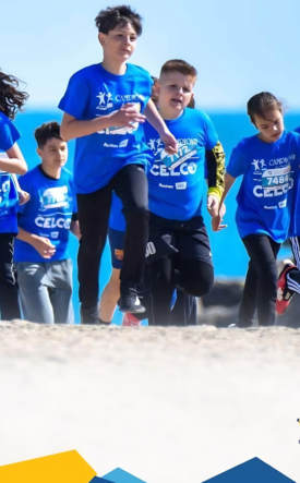 Group of children in blue shirts running on a beach during a marathon, with banners and logos visible in the foreground.