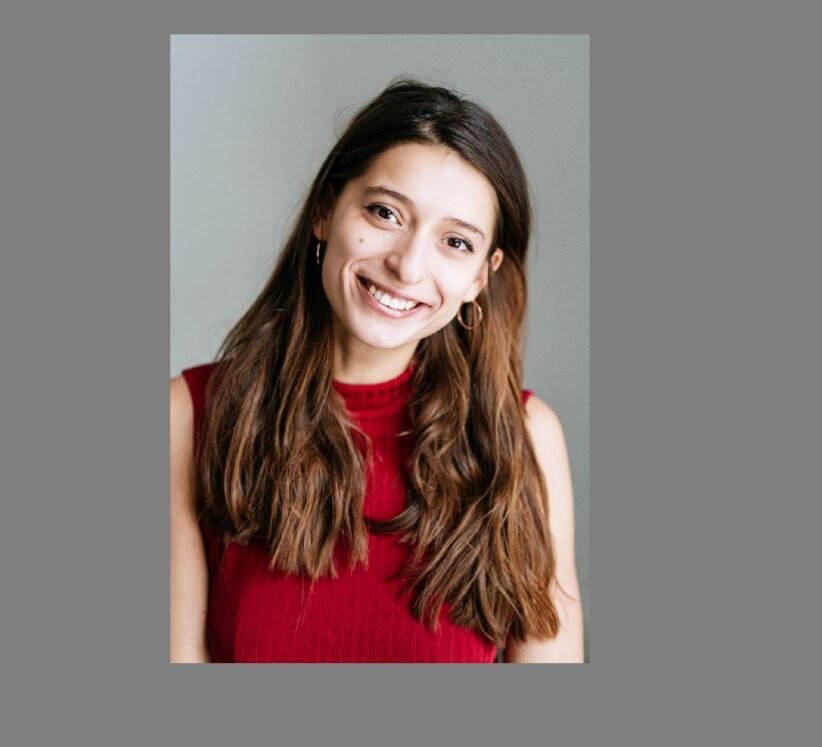 Smiling young woman with long brown hair wearing a red sleeveless top, posed against a neutral gray background.