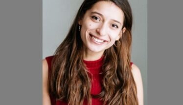 Smiling young woman with long brown hair wearing a red sleeveless top, posed against a neutral gray background.