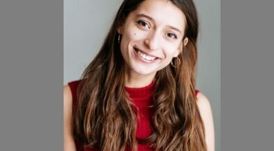 Smiling young woman with long brown hair wearing a red sleeveless top, posed against a neutral gray background.