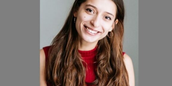 Smiling young woman with long brown hair wearing a red sleeveless top, posed against a neutral gray background.