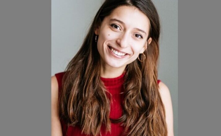 Smiling young woman with long brown hair wearing a red sleeveless top, posed against a neutral gray background.