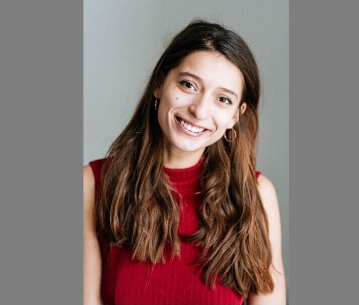 Smiling young woman with long brown hair wearing a red sleeveless top, posed against a neutral gray background.
