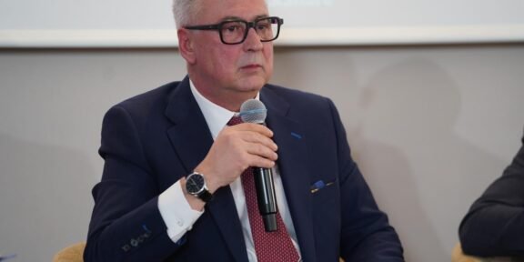 Man in a navy suit and red tie speaks into a handheld microphone at a panel discussion.