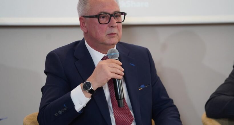 Man in a navy suit and red tie speaks into a handheld microphone at a panel discussion.