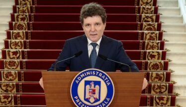 A man in a dark suit stands behind a wooden podium with a blue circular seal reading Administratia Prezidentială, speaking into two microphones, with red-carpeted stairs in the background.