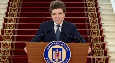 A man in a dark suit stands behind a wooden podium with a blue circular seal reading Administratia Prezidentială, speaking into two microphones, with red-carpeted stairs in the background.