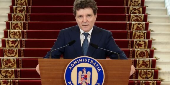 A man in a dark suit stands behind a wooden podium with a blue circular seal reading Administratia Prezidentială, speaking into two microphones, with red-carpeted stairs in the background.