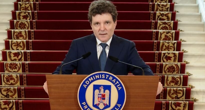 A man in a dark suit stands behind a wooden podium with a blue circular seal reading Administratia Prezidentială, speaking into two microphones, with red-carpeted stairs in the background.