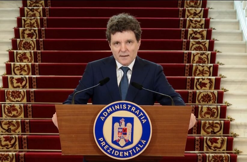 A man in a dark suit stands behind a wooden podium with a blue circular seal reading Administratia Prezidentială, speaking into two microphones, with red-carpeted stairs in the background.