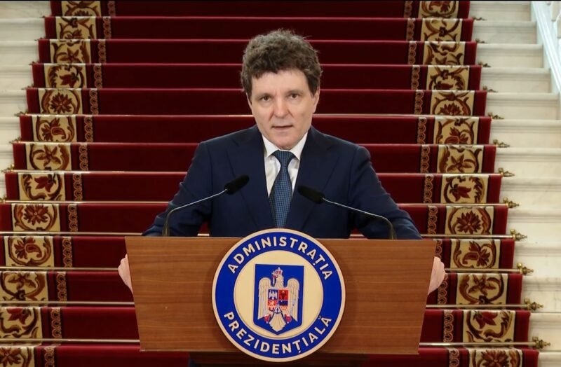 A man in a dark suit stands behind a wooden podium with a blue circular seal reading Administratia Prezidentială, speaking into two microphones, with red-carpeted stairs in the background.