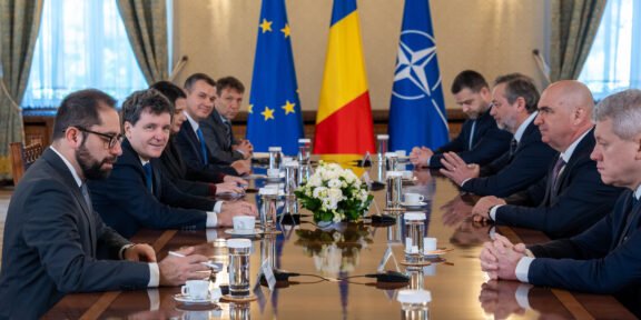 Diplomatic meeting at a long table with European Union, Romanian, and NATO flags behind, officials seated on both sides.