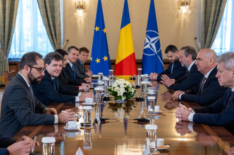 Diplomatic meeting at a long table with European Union, Romanian, and NATO flags behind, officials seated on both sides.