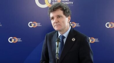 Man in a dark suit and blue tie speaks at a microphone during a press conference, with a blue backdrop featuring European Political Community logos.