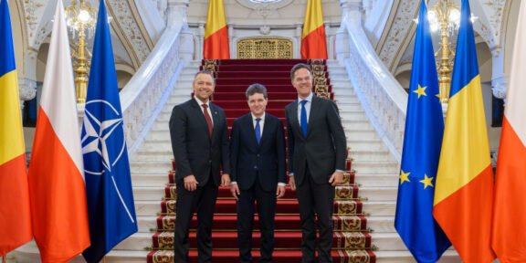 Three men in dark suits pose on a red-carpeted grand staircase, flanked by national flags including Romania, Poland, NATO, and the EU flag in a gilded hall.