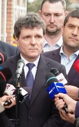 Man in a dark suit giving a press statement, surrounded by reporters and microphones outdoors.