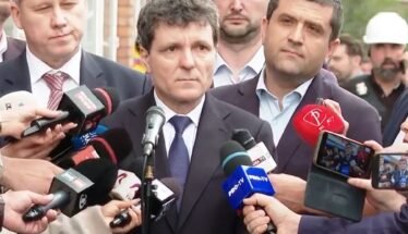 Man in a dark suit giving a press statement, surrounded by reporters and microphones outdoors.