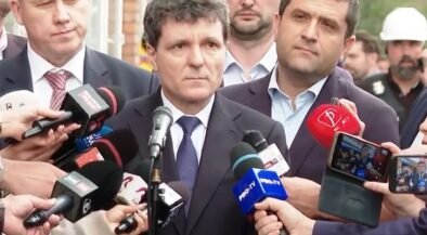 Man in a dark suit giving a press statement, surrounded by reporters and microphones outdoors.