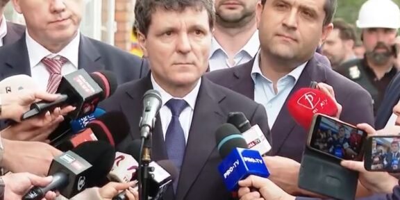 Man in a dark suit giving a press statement, surrounded by reporters and microphones outdoors.