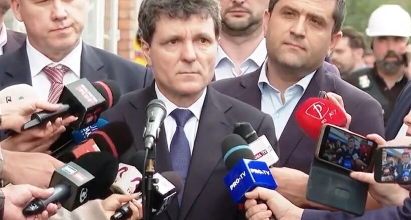 Man in a dark suit giving a press statement, surrounded by reporters and microphones outdoors.