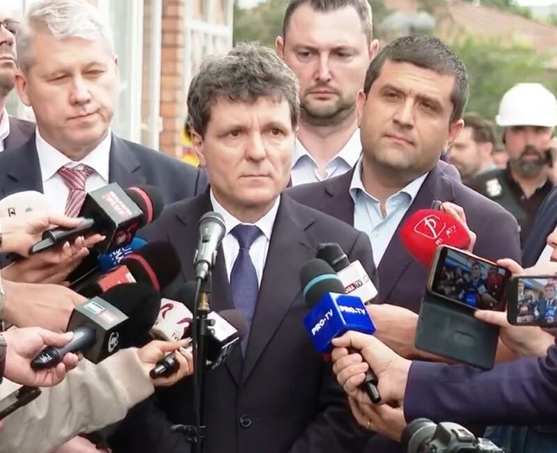 Man in a dark suit giving a press statement, surrounded by reporters and microphones outdoors.