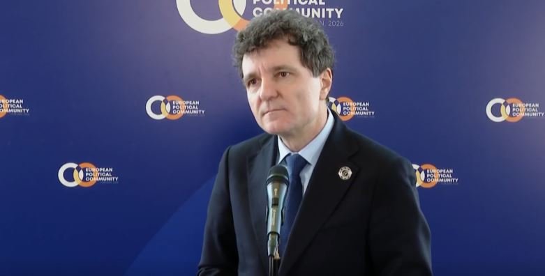 Man in a dark suit and blue tie speaks at a microphone during a press conference, with a blue backdrop featuring European Political Community logos.