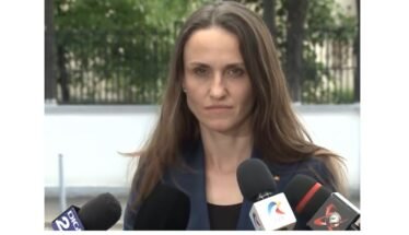 Woman in a navy blazer speaks to reporters at an outdoor press conference, multiple microphones in front of her.