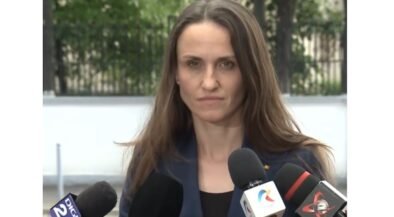 Woman in a navy blazer speaks to reporters at an outdoor press conference, multiple microphones in front of her.