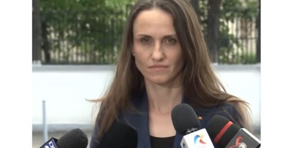 Woman in a navy blazer speaks to reporters at an outdoor press conference, multiple microphones in front of her.
