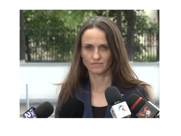 Woman in a navy blazer speaks to reporters at an outdoor press conference, multiple microphones in front of her.