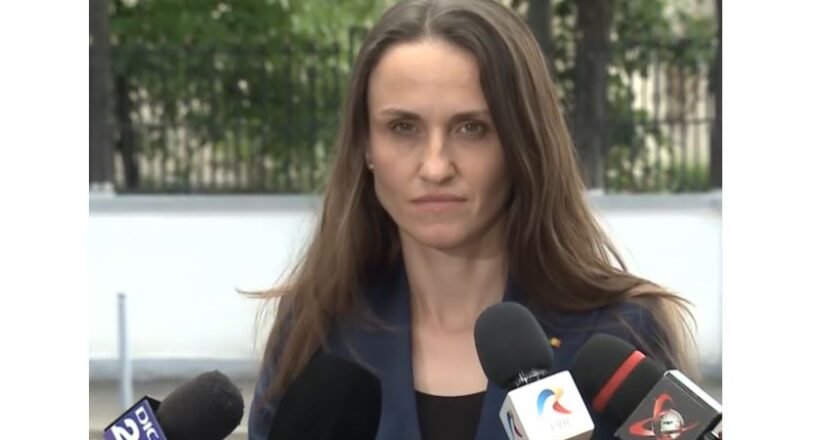 Woman in a navy blazer speaks to reporters at an outdoor press conference, multiple microphones in front of her.