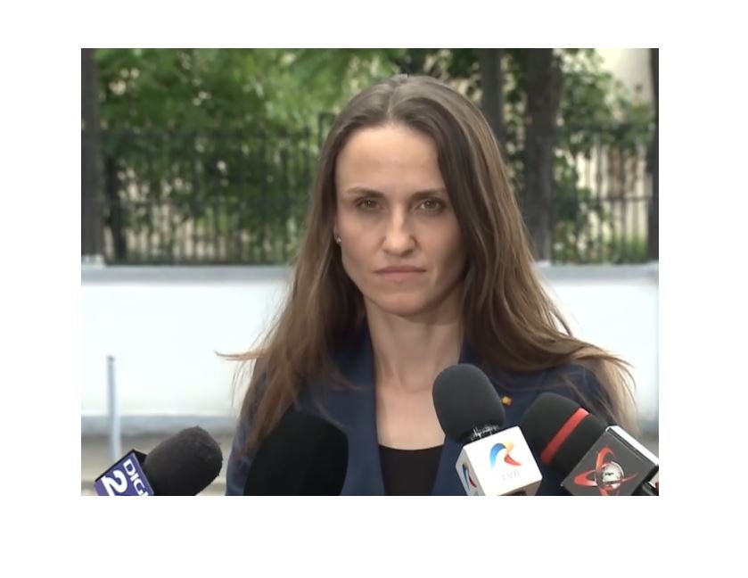 Woman in a navy blazer speaks to reporters at an outdoor press conference, multiple microphones in front of her.