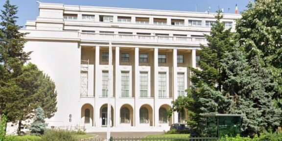 White neoclassical building with tall columns, arches at ground level, gated lawn, and surrounding trees.