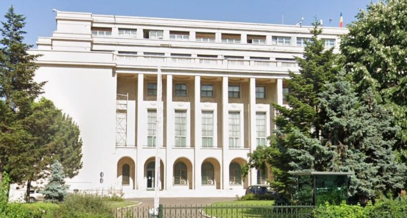 White neoclassical building with tall columns, arches at ground level, gated lawn, and surrounding trees.
