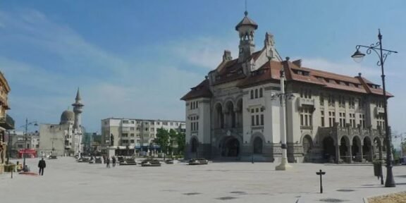 Historic building with arches on the ground floor and a steep red roof, in a sunlit urban square.
