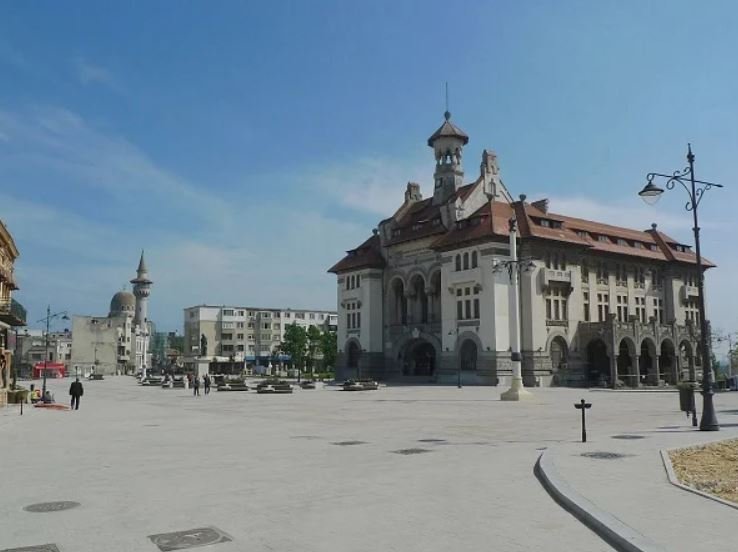Historic building with arches on the ground floor and a steep red roof, in a sunlit urban square.