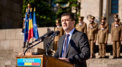 Man in a dark suit delivering a speech at a wooden podium labeled ROMANIA, with flags and soldiers in ceremonial uniforms behind him.