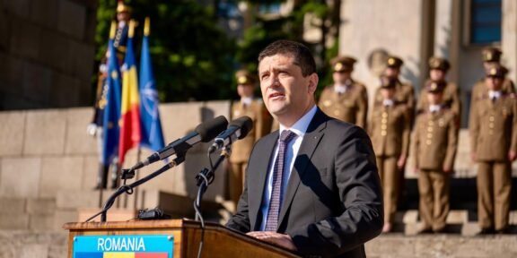 Man in a dark suit delivering a speech at a wooden podium labeled ROMANIA, with flags and soldiers in ceremonial uniforms behind him.
