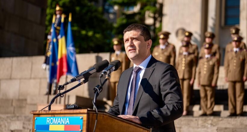 Man in a dark suit delivering a speech at a wooden podium labeled ROMANIA, with flags and soldiers in ceremonial uniforms behind him.