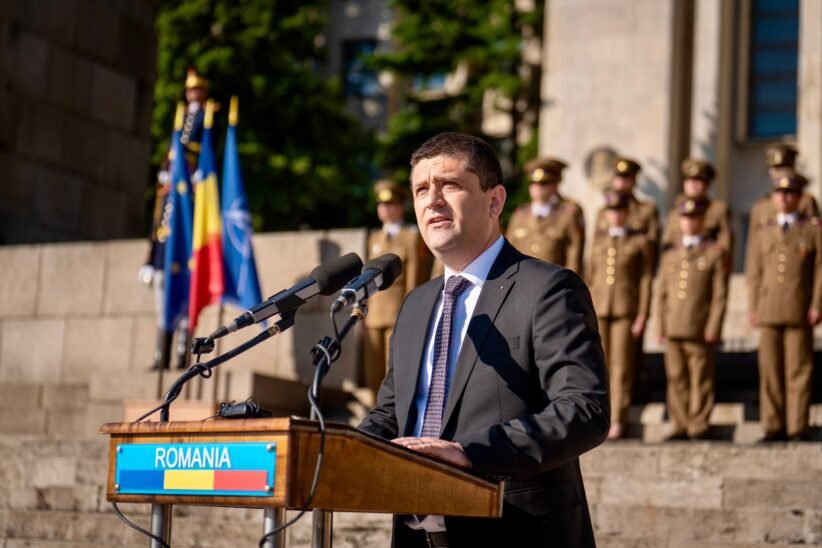 Man in a dark suit delivering a speech at a wooden podium labeled ROMANIA, with flags and soldiers in ceremonial uniforms behind him.
