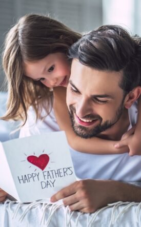 Father and daughter cuddling on a bed, smiling at a card that reads 'Happy Father's Day' with a red heart.