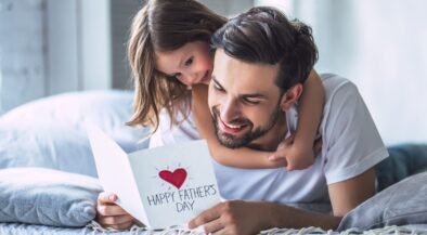 Father and daughter cuddling on a bed, smiling at a card that reads 'Happy Father's Day' with a red heart.