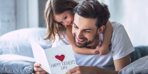 Father and daughter cuddling on a bed, smiling at a card that reads 'Happy Father's Day' with a red heart.