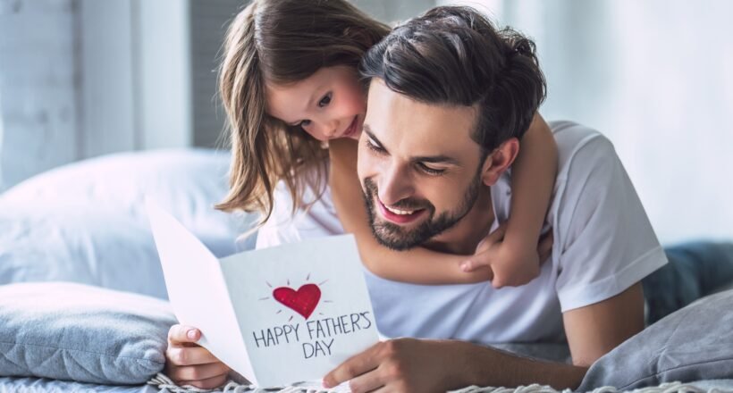 Father and daughter cuddling on a bed, smiling at a card that reads 'Happy Father's Day' with a red heart.