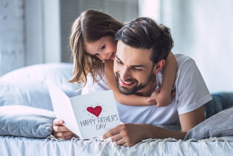 Father and daughter cuddling on a bed, smiling at a card that reads 'Happy Father's Day' with a red heart.
