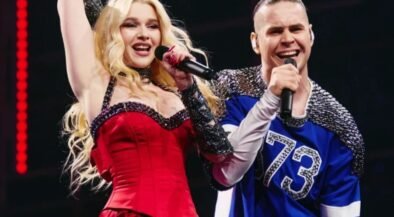 Two performers sing on stage: a blonde woman in a red dress raising her arm, holding a microphone, beside a man in a blue jersey with a mic.