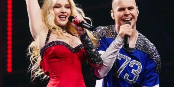 Two performers sing on stage: a blonde woman in a red dress raising her arm, holding a microphone, beside a man in a blue jersey with a mic.