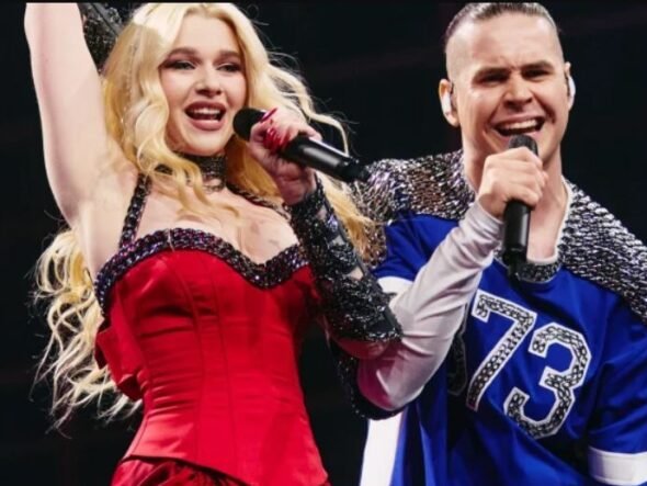 Two performers sing on stage: a blonde woman in a red dress raising her arm, holding a microphone, beside a man in a blue jersey with a mic.
