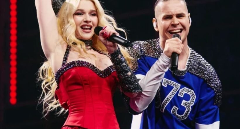 Two performers sing on stage: a blonde woman in a red dress raising her arm, holding a microphone, beside a man in a blue jersey with a mic.
