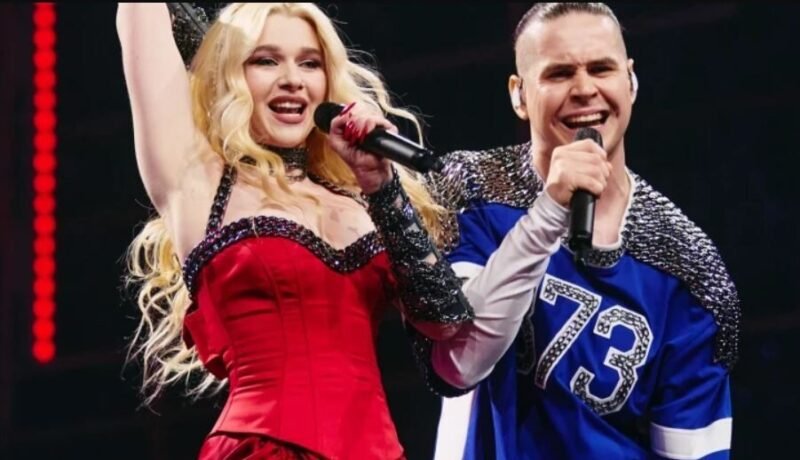 Two performers sing on stage: a blonde woman in a red dress raising her arm, holding a microphone, beside a man in a blue jersey with a mic.